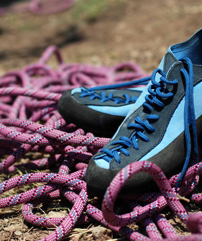 Zapatos de escalada en primer plano con cordones azules sobre un fondo de cuerda de escalada rosa y violeta, situados en un suelo natural de tierra. La imagen evoca la actividad al aire libre y la aventura, perfecta para destacar la necesidad del protector solar &